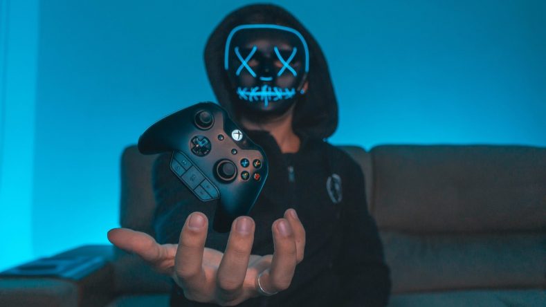 A masked individual holding an Xbox controller with a vibrant blue background.